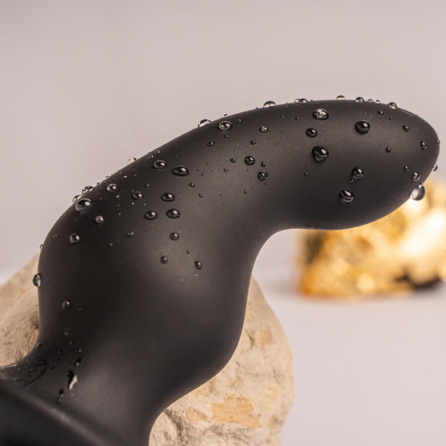 Macro de la tige courbe à picots du plug noir couverte de gouttes d'eau, calée près d'une pierre claire, silicone lisse illustrant l'étanchéité de l'accessoire.
