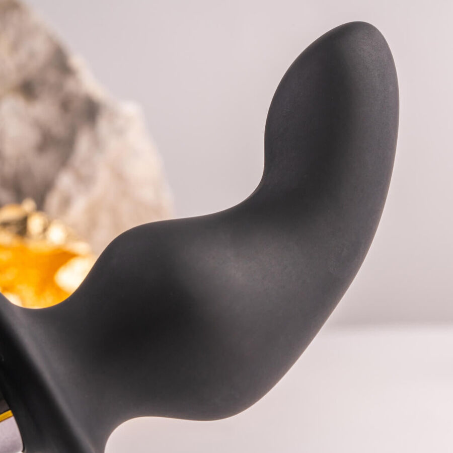 Tige souple recourbée du plug noir dressée près d'une grosse pierre claire, courbe lisse en silicone sur fond neutre, sans aucun détail anatomique ni relief réaliste.