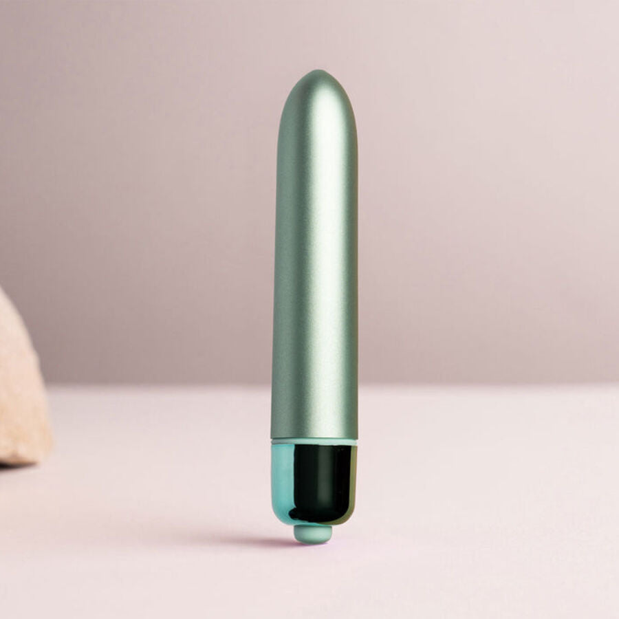 Mini bullet vert dressé debout sur surface rose pâle, capuchon de commande noir bien visible, corps fin et lisse devant un arrière-plan flouté neutre et discret.