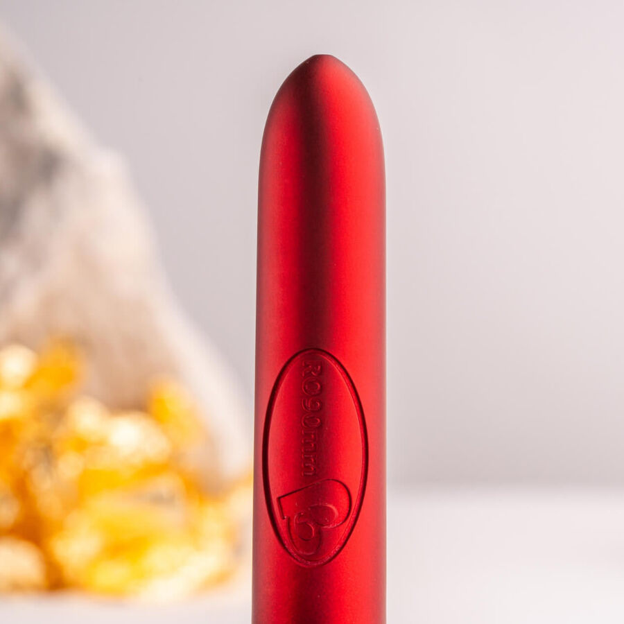 Bullet rouge satiné dressé près d'une pierre claire et d'un éclat doré, logo Rocks-Off gravé sur le corps, surface lisse métallisée sur fond beige neutre et sobre.