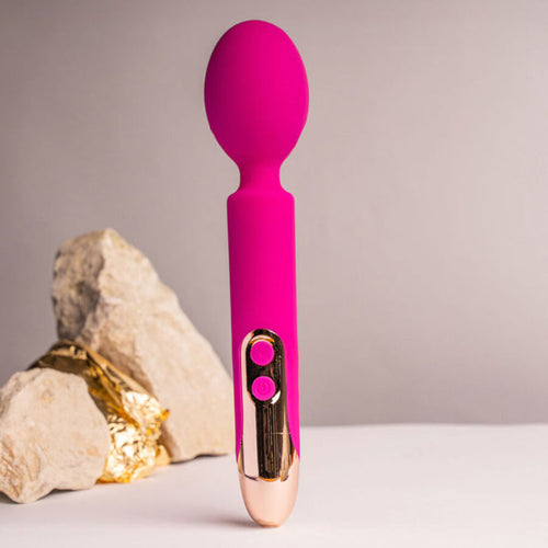 Mini wand rose à grosse tête souple dressé près de pierres claires, manche orné de deux boutons ronds, finition silicone lisse et brillante sur fond neutre et épuré.