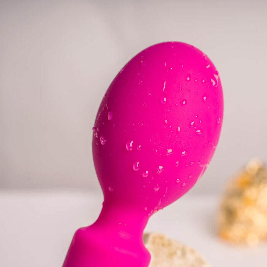 Gros plan de la tête bulbeuse rose du wand couverte de gouttes d'eau, manche silicone lisse, éclat doré flouté en arrière-plan soulignant l'étanchéité.