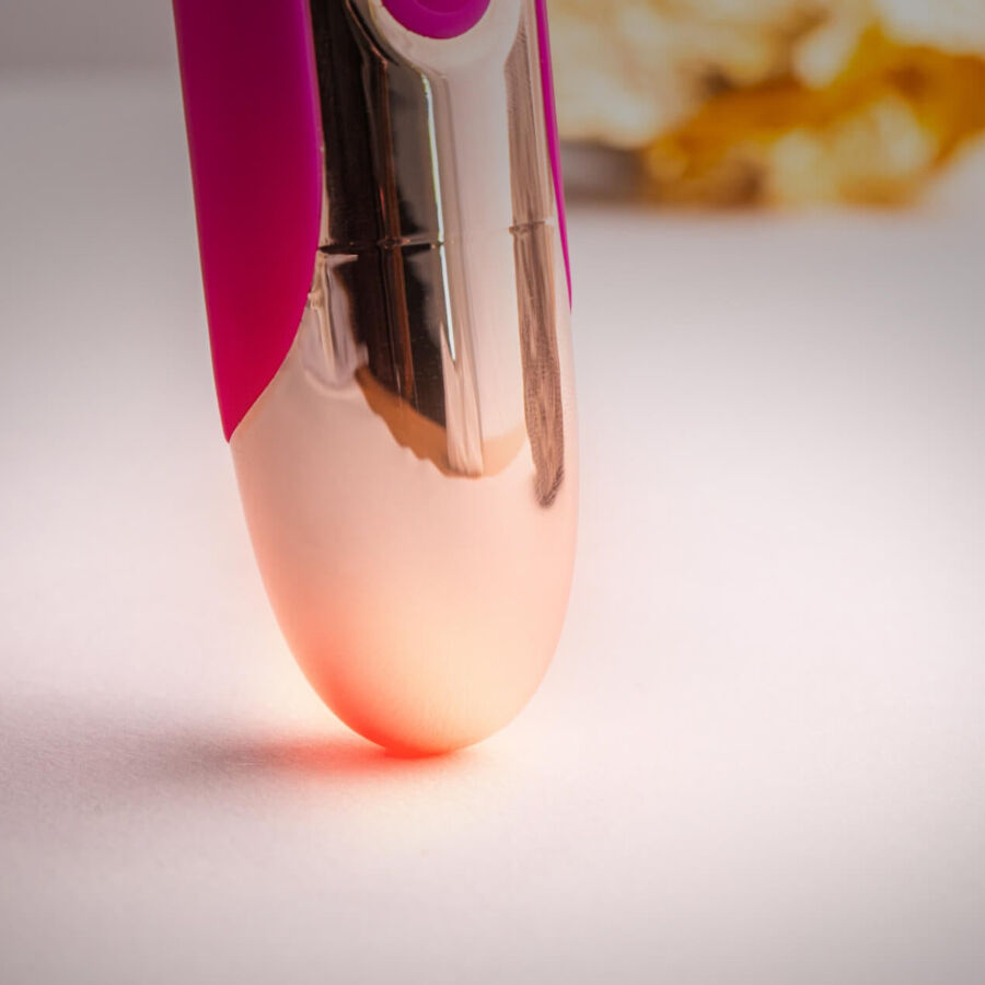 Gros plan de la base dorée brillante du wand rose en contre-plongée, jonction métal et silicone éclairée d'une lueur chaude, surface lisse réfléchissante.