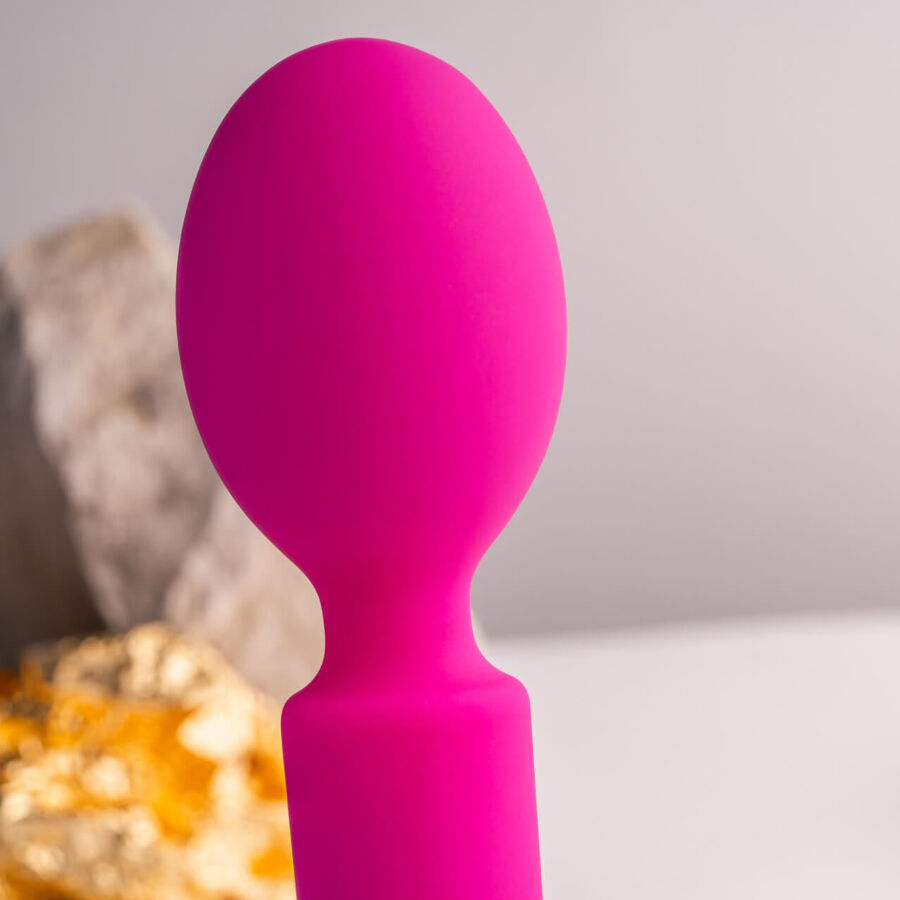 Macro de la tête souple arrondie du wand rose et de son col fin, posée près d'une pierre claire, silicone lisse mis en valeur par un éclairage doux et chaleureux.