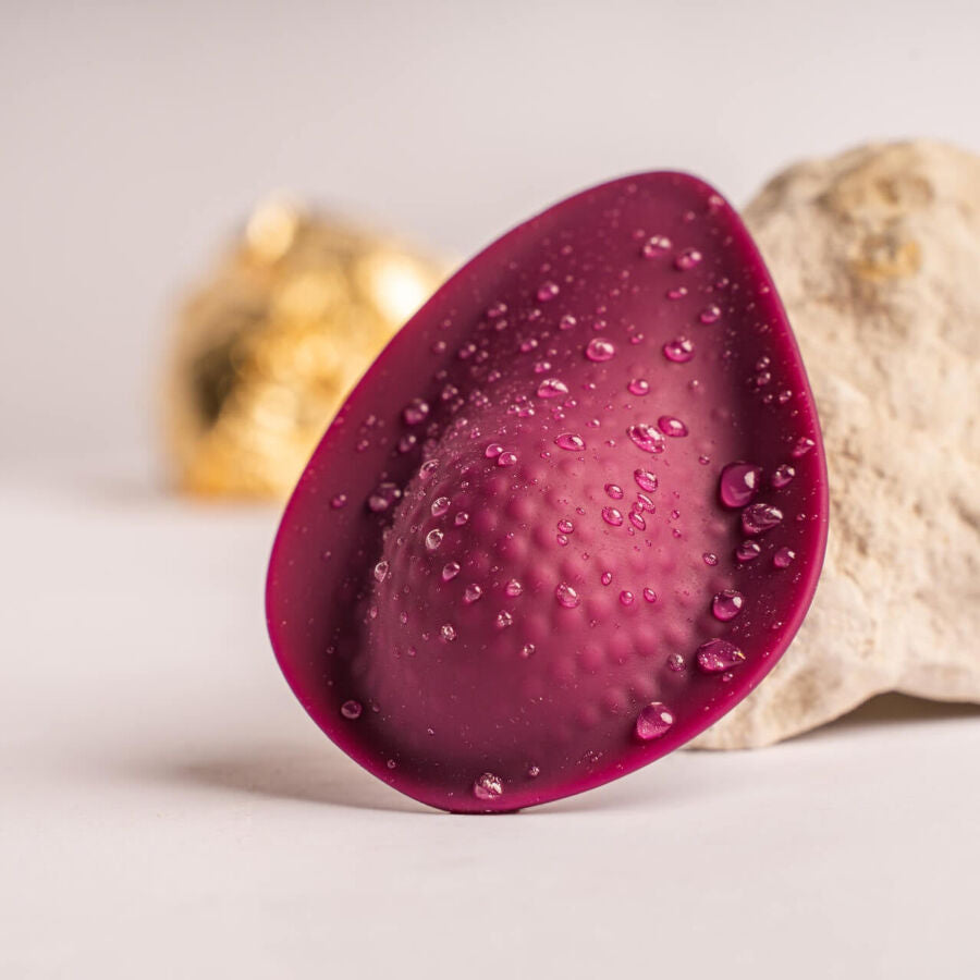Stimulateur bordeaux en forme de goutte couvert de gouttes d'eau, posé près d'une pierre claire, picots de la face interne visibles, silicone lisse et entièrement étanche.