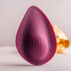 Vue de profil du stimulateur intime bordeaux en goutte, face picotée orientée vers l'avant, éclat doré flouté en fond, surface silicone douce et lisse sur fond clair.