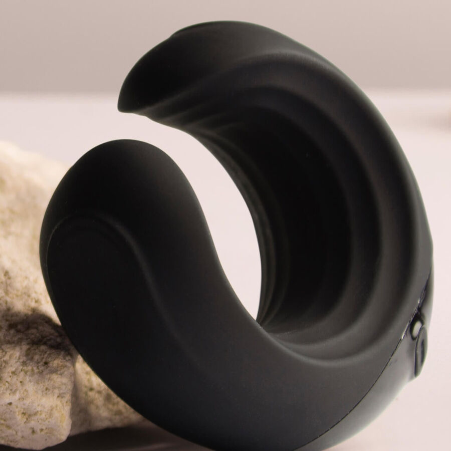 Anneau pénien noir en croissant calé entre deux pierres claires, surface silicone lisse et logement de la bullet vibrante apparent à la base, sur fond beige sobre.