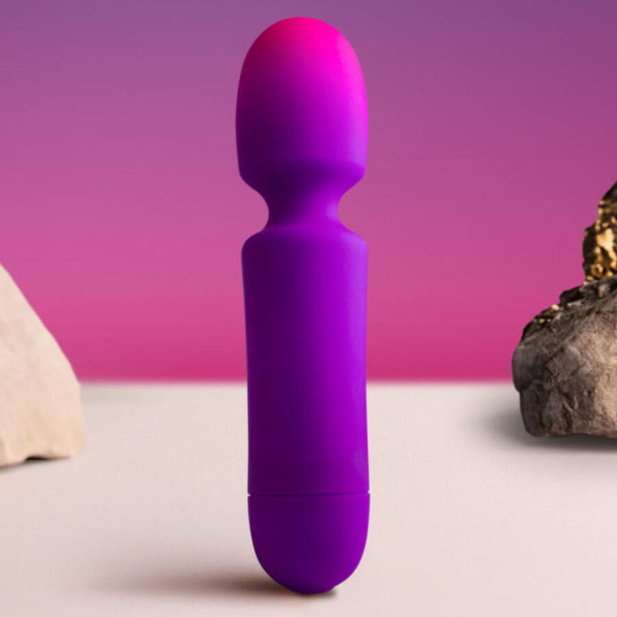 Mini wand vibrant rose-violet debout sur fond rosé près de pierres, tête boule flexible et manche galbé visibles, format compact en silicone lisse non anatomique.