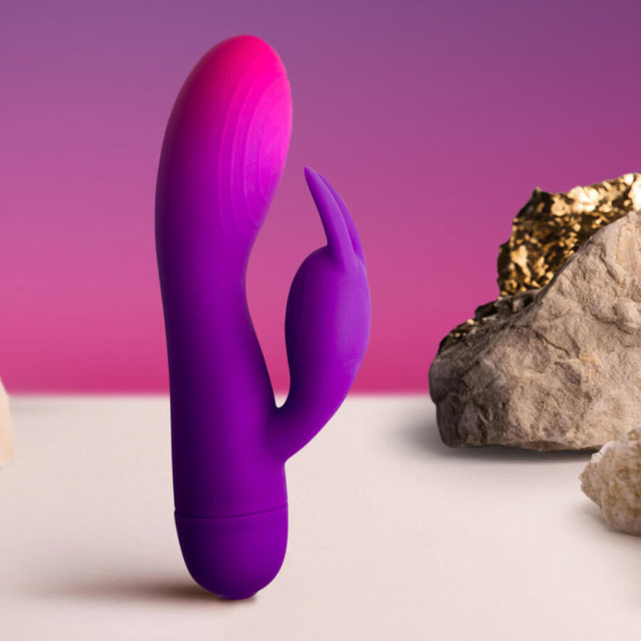 Vibromasseur rabbit double moteur dressé de profil sur fond rose, longue tige courbe et oreille clitoridienne fine, silicone lisse mis en valeur près d'un caillou.