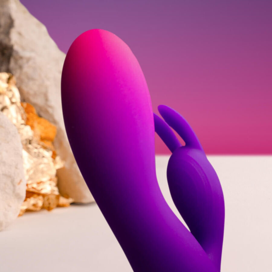 Gros plan du rabbit silicone rose-violet posé devant des galets clairs, tige bombée et bras clitoridien souple visibles, finition veloutée lisse sans détail anatomique.