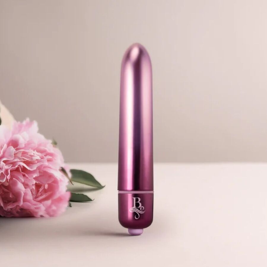 Bullet vibrant violet métallisé dressé devant des fleurs roses, embout coloré visible, mini vibromasseur clitoridien compact à finition brillante Rocks-Off.