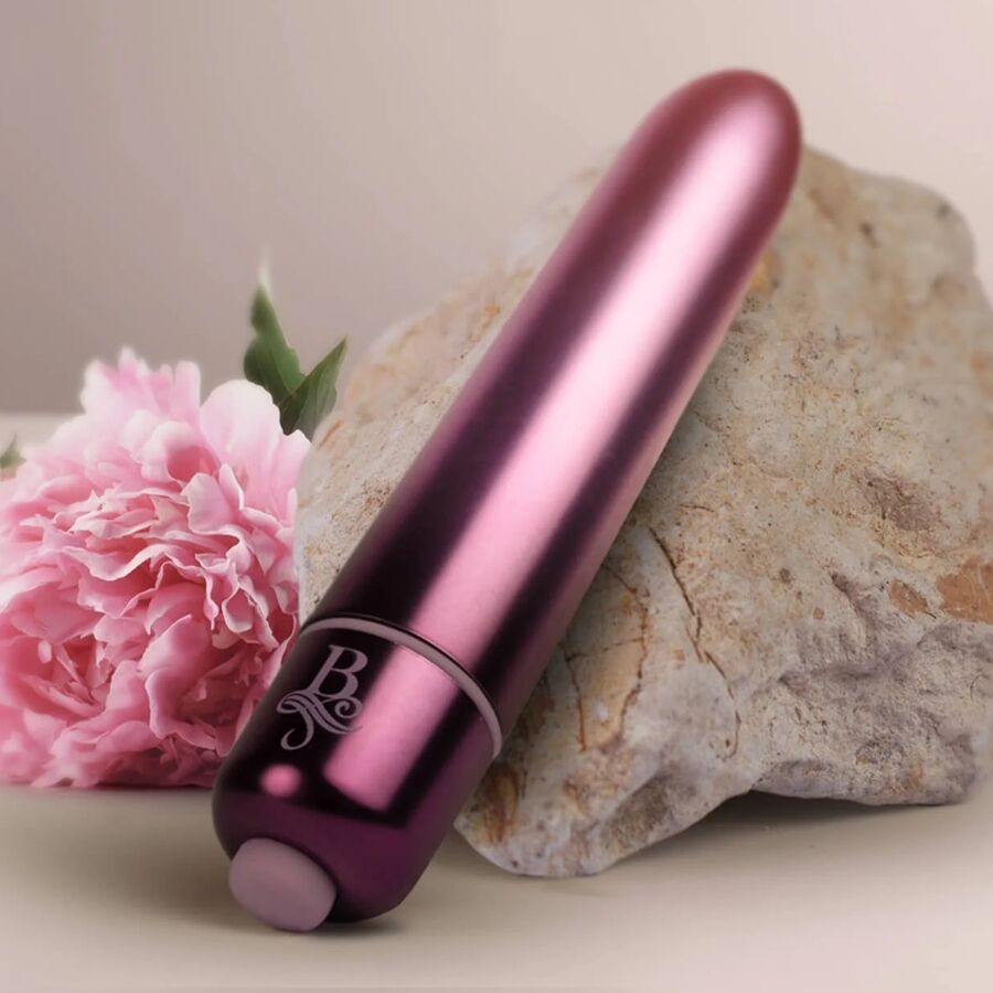 Bullet vibrant bordeaux métallisé posé contre une pierre avec une fleur rose, embout de commande visible, mini vibromasseur clitoridien compact à finition brillante.