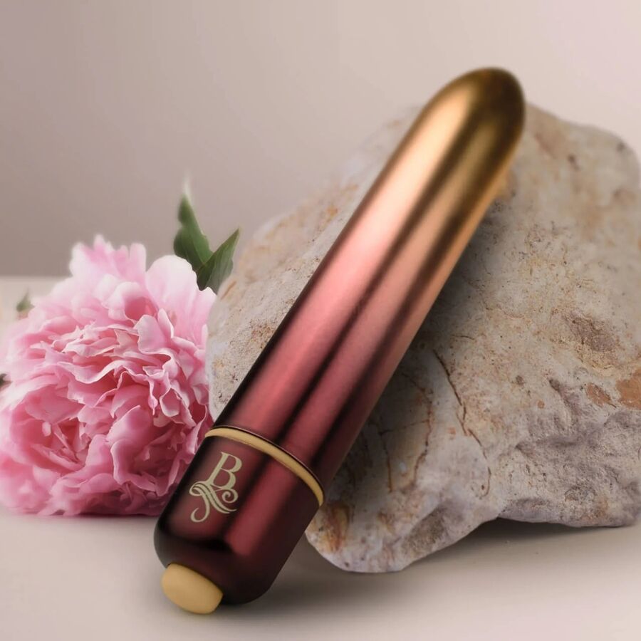 Bullet vibrant dégradé or-rouge posé contre une pierre avec une fleur rose, embout doré visible, mini vibromasseur clitoridien compact à finition métallisée bicolore.