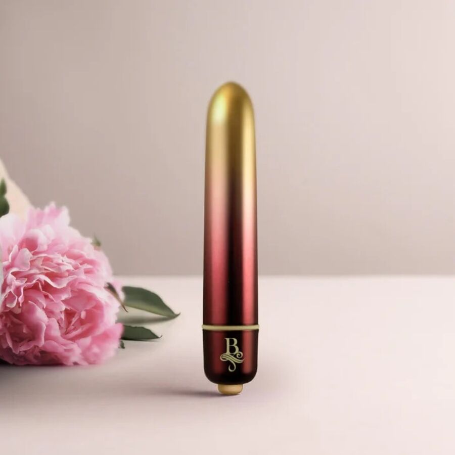 Bullet vibrant dégradé or-rouge dressé devant des fleurs roses, pointe arrondie, mini vibromasseur clitoridien compact à finition métallisée bicolore Rocks-Off.