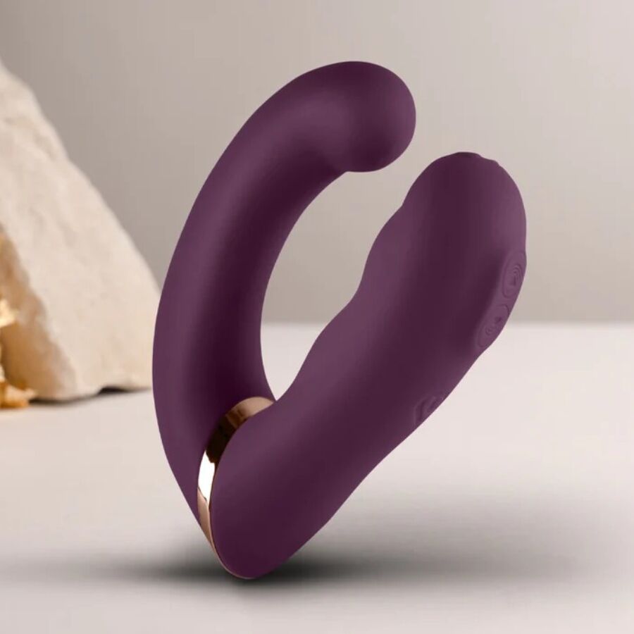 Double vibromasseur violet en U posé près d'une pierre claire, deux bras incurvés et liseré doré visibles, silicone mat lisse non anatomique, mise en scène.