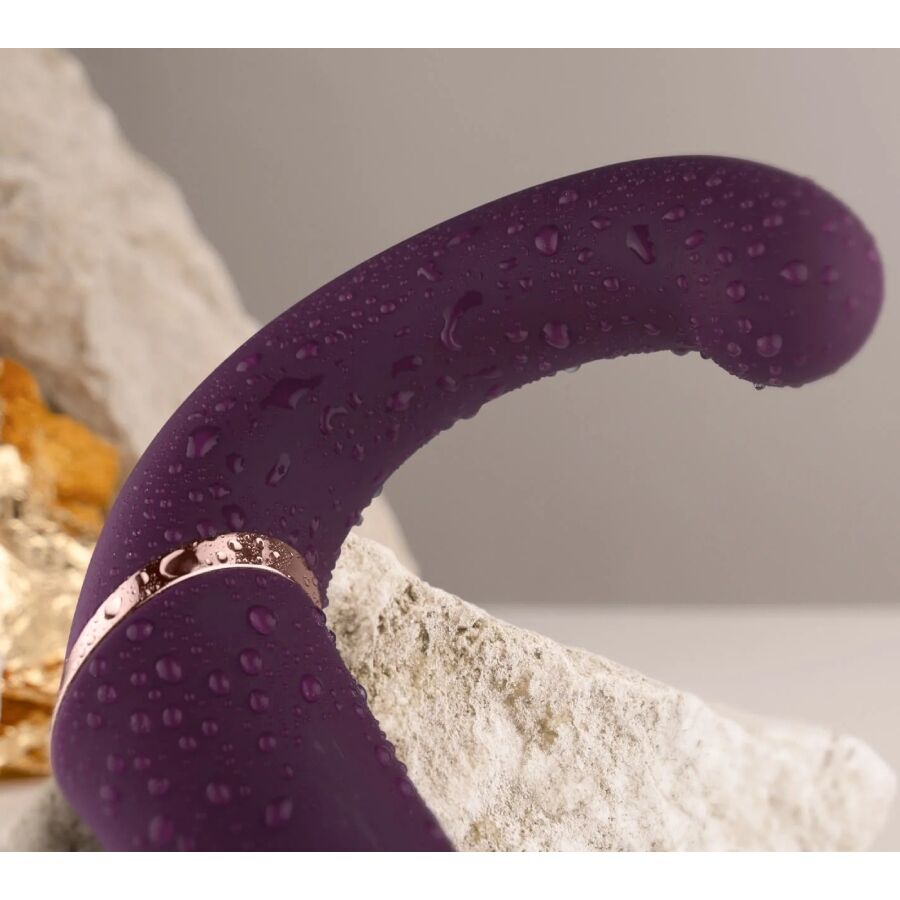 Gros plan d'un bras texturé du double vibromasseur violet perlé de gouttes d'eau près d'une pierre, liseré doré visible, illustrant l'étanchéité du jouet.