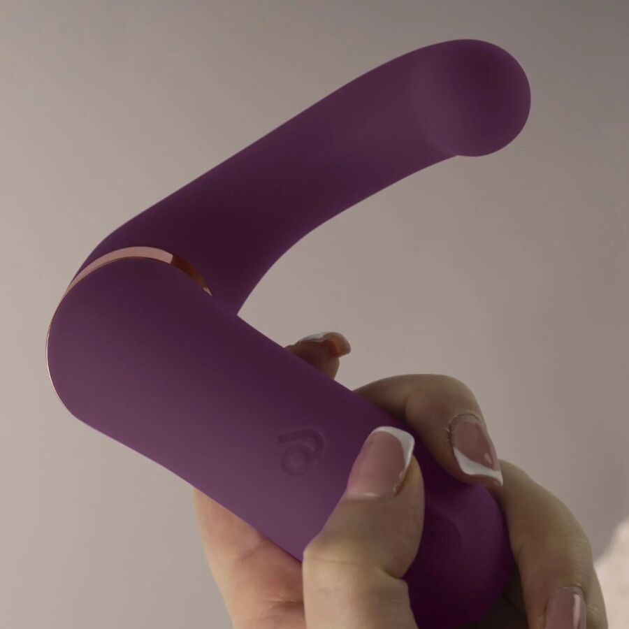 Main féminine tenant le double vibromasseur violet en U par sa base, montrant la flexibilité des deux bras et le liseré doré du jouet en silicone non anatomique.