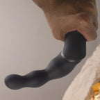 Stimulateur prostatique noir à double bras en gros plan contre un décor minéral, embout arrondi et courbe flexible visibles, silicone doux non anatomique.