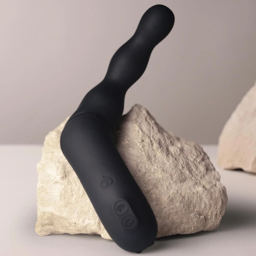 Stimulateur prostatique noir à double bras posé contre des pierres claires, boutons de commande visibles sur le corps, silicone mat lisse non anatomique.
