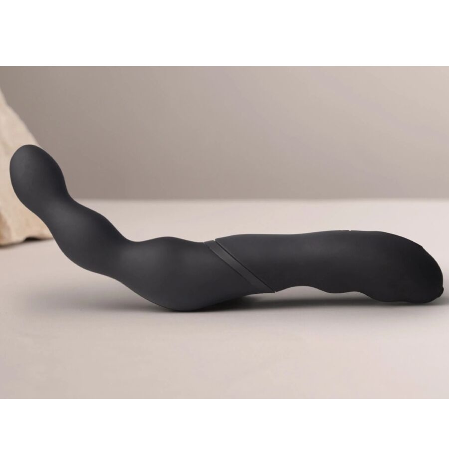 Stimulateur prostatique noir à double bras posé à plat sur fond beige, profil bas et embouts arrondis visibles, surface silicone mate lisse non anatomique.