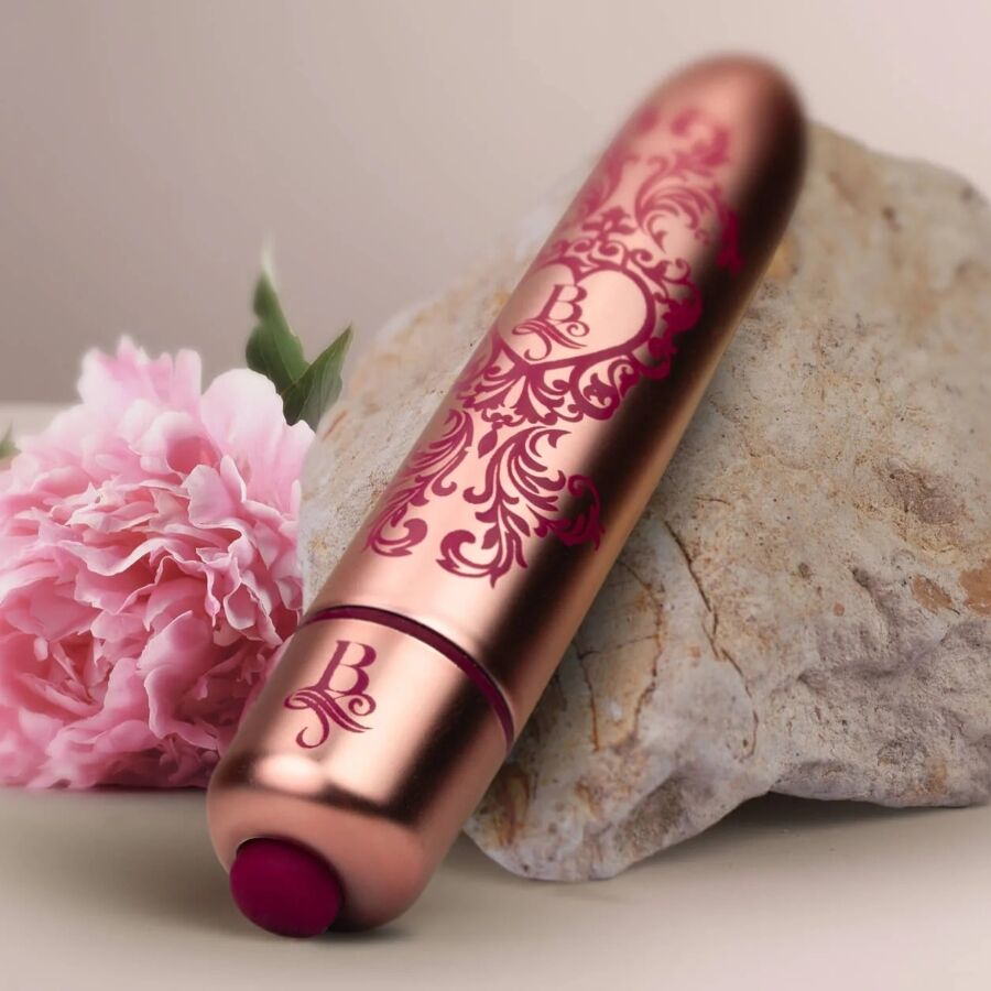 Bullet vibrant rose doré gravé posé contre une pierre avec une fleur rose, embout bordeaux visible, mini vibromasseur clitoridien à finition baroque métallisée.