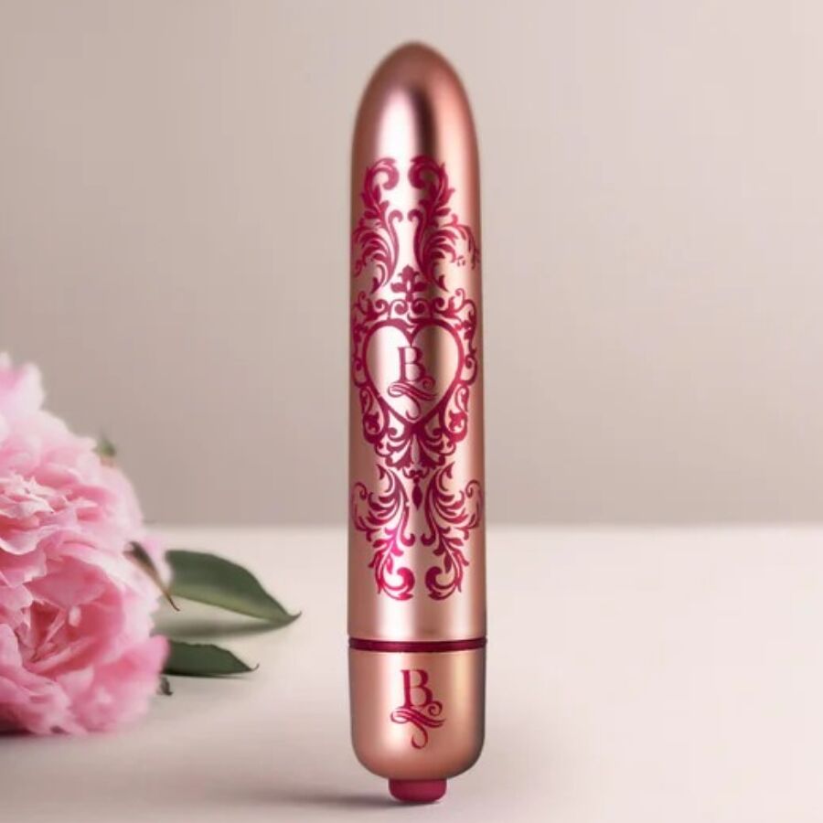 Bullet vibrant rose doré gravé posé devant des fleurs roses, mini vibromasseur clitoridien fin à la finition baroque métallisée et motifs bordeaux Rocks-Off.