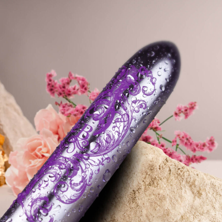 Gros plan du bullet vibrant violet gravé couvert de gouttes d'eau devant des fleurs, finition baroque illustrant l'étanchéité de ce mini vibromasseur clitoridien.