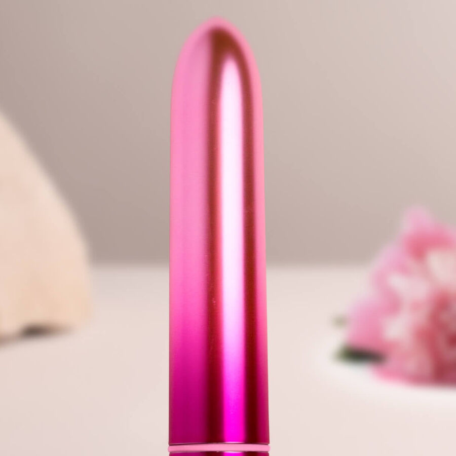 Bullet vibrant rose vif dressé sur sa base devant un décor flou de fleurs, pointe arrondie, mini vibromasseur clitoridien compact à finition métallisée brillante.