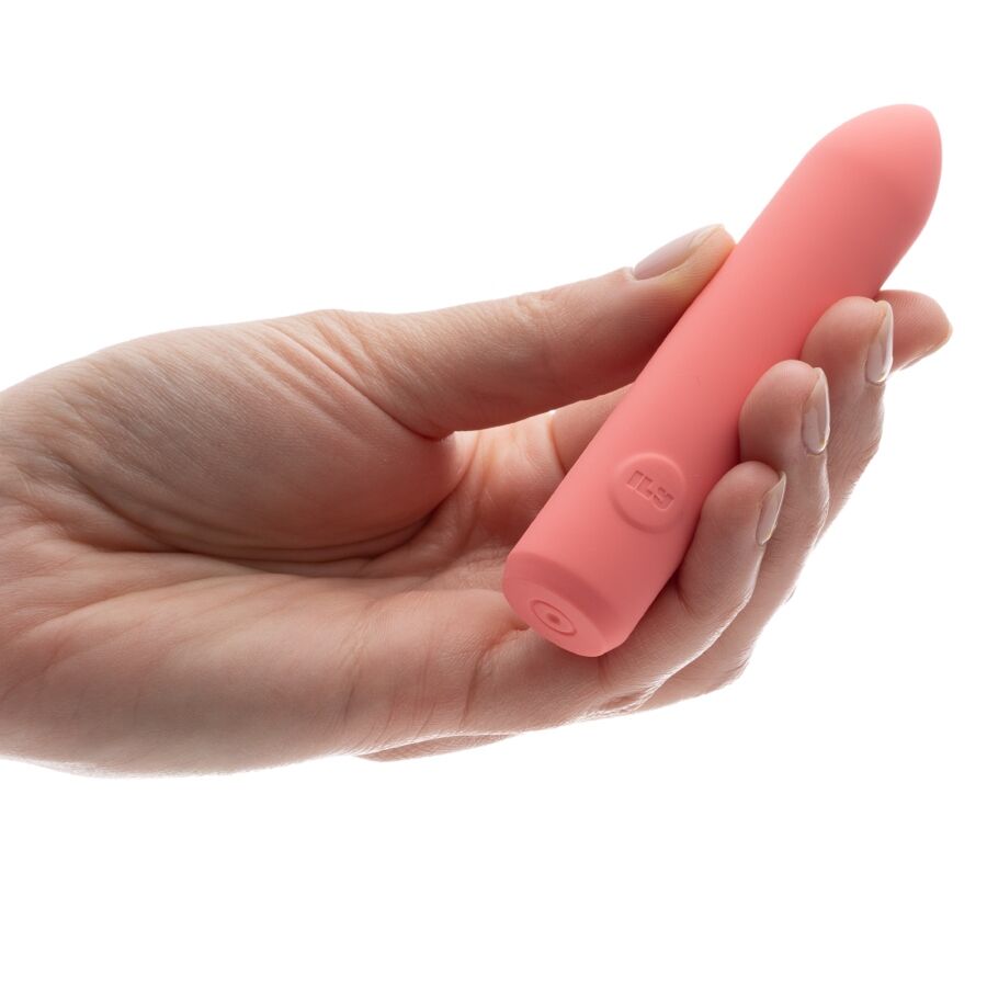 Bullet corail tenu dans une main aux ongles clairs, cylindre lisse à pointe arrondie dépassant entre les doigts, plan de prise rapproché du produit sur fond blanc parfaitement épuré.