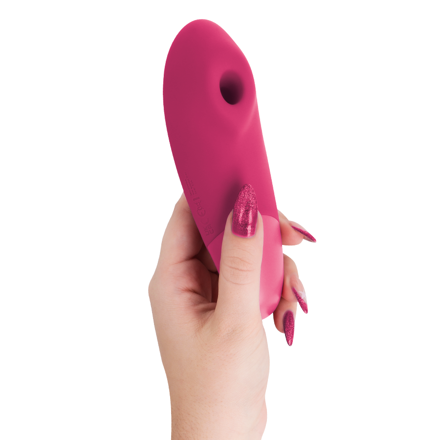 Main aux ongles framboise tenant le stimulateur clitoridien framboise Womanizer Enhance de profil, embout de succion vers le haut, corps galbé lisse en silicone.