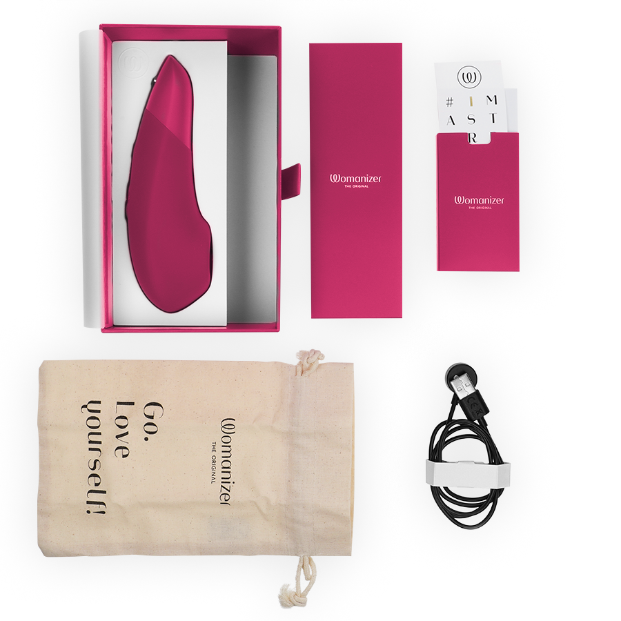 Coffret Womanizer Enhance ouvert : stimulateur framboise dans l'écrin, pochette 'Go love yourself', notice et câble de charge magnétique, disposés à plat fond blanc.