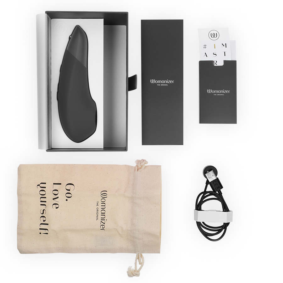 Coffret Womanizer Enhance ouvert : stimulateur noir dans l'écrin, pochette 'Go love yourself', notice et câble de charge magnétique, disposés à plat sur fond blanc.