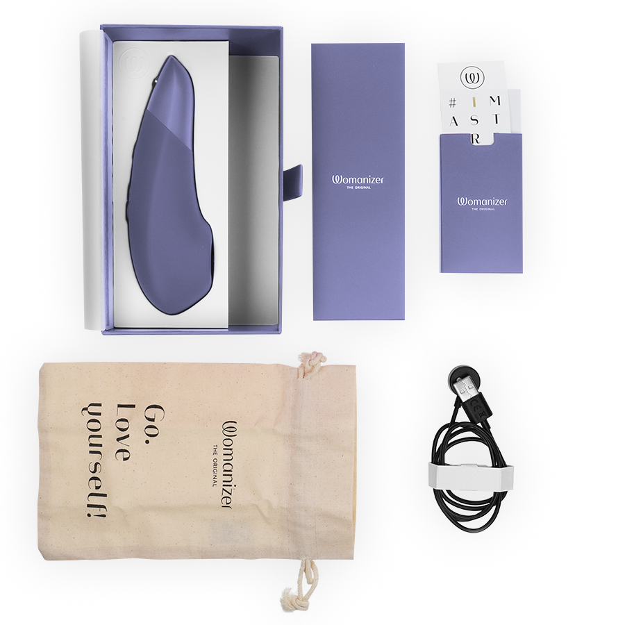 Coffret Womanizer Enhance ouvert : stimulateur lilas dans l'écrin, pochette 'Go love yourself', notice et câble de charge magnétique, disposés à plat sur fond blanc.