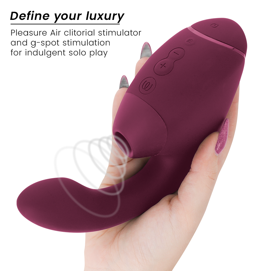 Main aux ongles violets tenant le stimulateur double bordeaux Womanizer de face : embout d'air pulsé clitoridien et tige interne vibrante courbée en silicone doux.