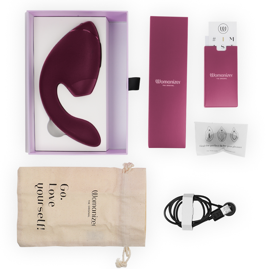 Coffret Womanizer ouvert avec le stimulateur double bordeaux dans l'écrin, embouts, pochette 'Go love yourself', notice et câble magnétique, disposés à plat.