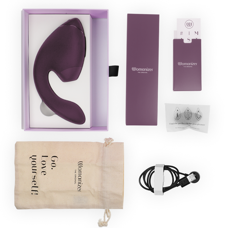 Coffret Womanizer ouvert avec le stimulateur double prune dans l'écrin, embouts de rechange, pochette 'Go love yourself' et câble de charge magnétique, vue à plat.