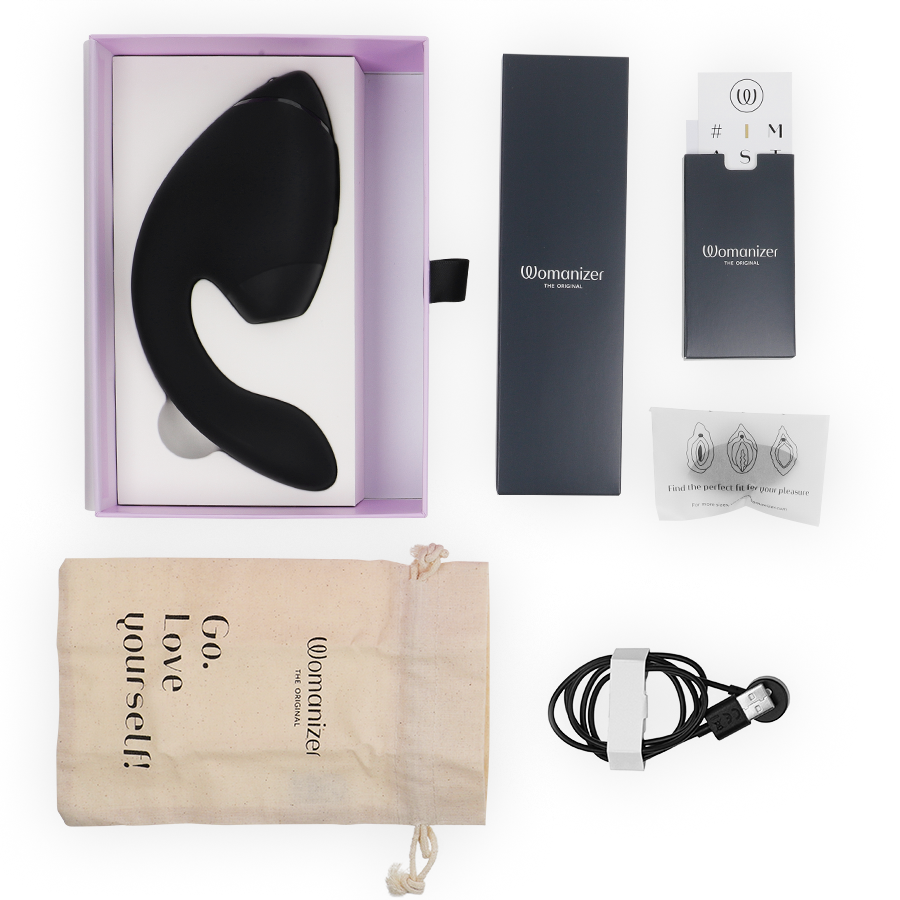 Coffret Womanizer ouvert : stimulateur double noir dans l'écrin, pochette 'Go love yourself', notice, câble de charge magnétique et embouts, disposés à plat.