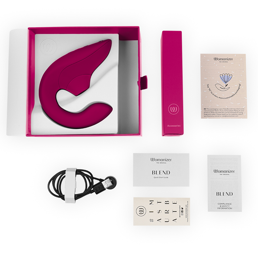 Coffret Womanizer ouvert montrant le stimulateur rose dans son écrin, notice pliée, câble de charge USB magnétique et sachet, présentés à plat sur fond blanc.