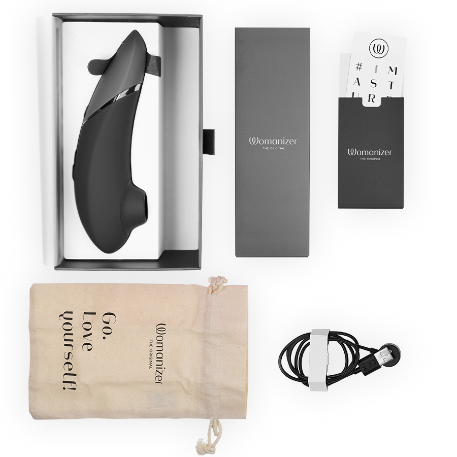 Womanizer Next noir présenté avec son écrin, câble de charge magnétique, pochette Go love yourself et cartes d'accessoires disposés sur fond blanc épuré.