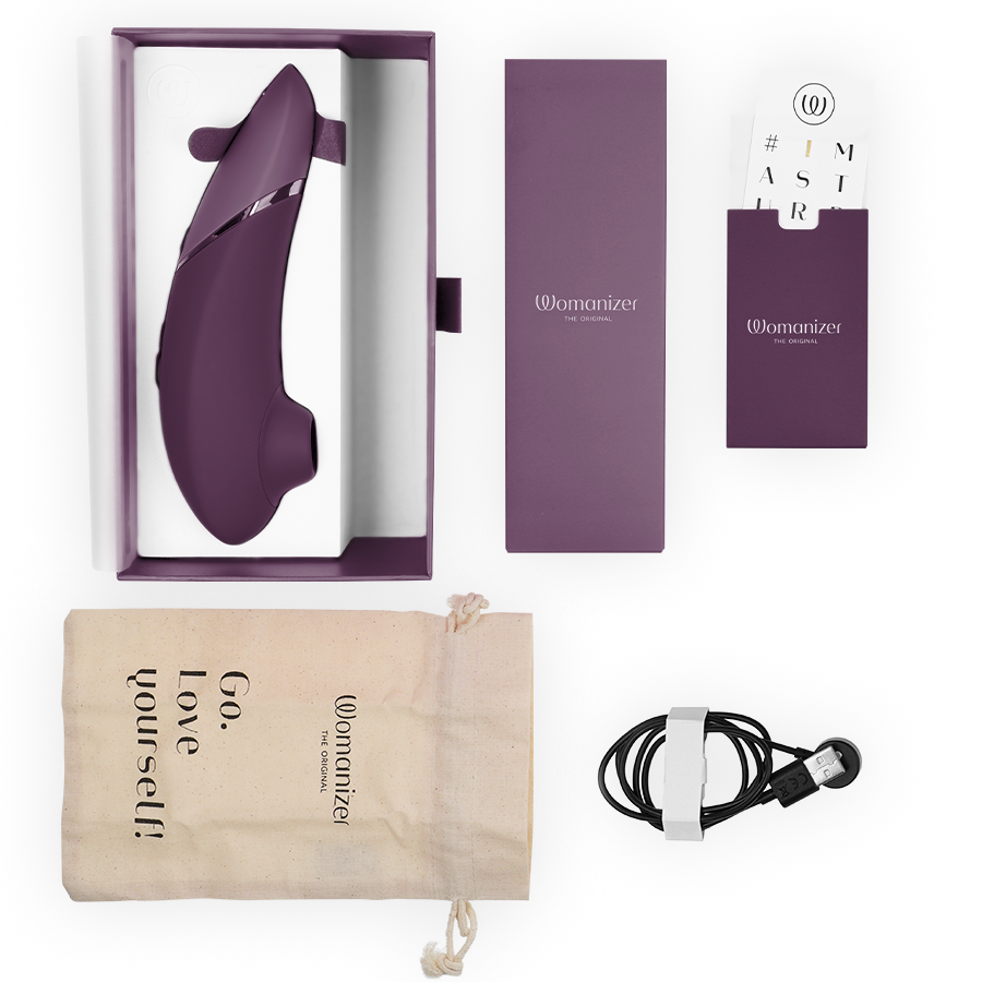 Womanizer Next aubergine présenté avec son écrin prune, câble de charge magnétique, pochette Go love yourself et cartes d'accessoires disposés sur fond clair.
