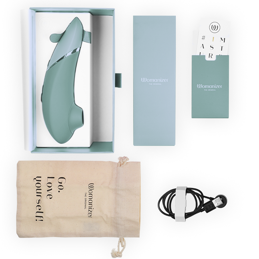 Womanizer Next vert sauge présenté avec son écrin clair, câble de charge magnétique, pochette Go love yourself et cartes d'accessoires disposés sur fond blanc.