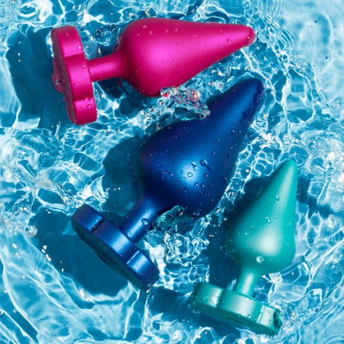 Plugs anaux ROMP multicolores metallises en silicone poses sur fond aquatique, formes coniques lisses et bases fleur, kit progressif etanche et stable.