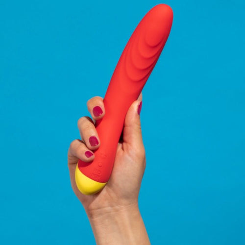 Main tenant le vibromasseur point G ROMP corail sur fond bleu, montrant la taille elancee et la tete incurvee du jouet en silicone soyeux.