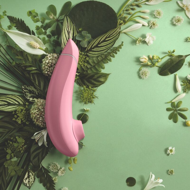 Womanizer Premium Eco rose posé parmi des feuilles et des fleurs sur un fond vert.