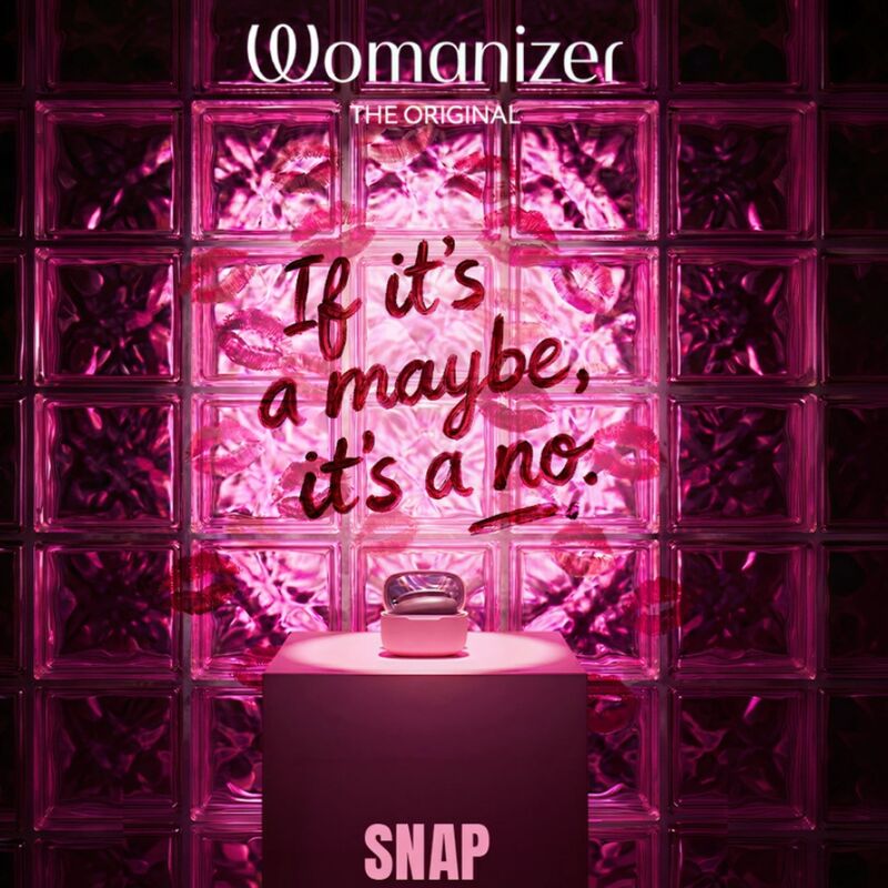 Étui rose du Womanizer Snap ouvert avec le stimulateur Baie écrasée à l’intérieur, devant des blocs de verre.