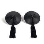 Paire de cache-tétons noirs à pompons et nœud satin vus de face, accessoire de poitrine burlesque adhésif et réutilisable Bijoux Indiscrets pour le décolleté.