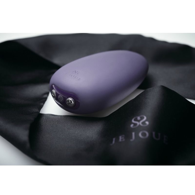 Vibrateur Mimi Soft violet posé sur un tissu satiné noir griffé Je Joue, galet en silicone mat mis en scène, ambiance feutrée et raffinée sans nudité.