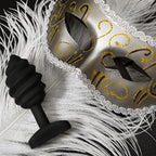 Plug anal vibrant G-Vibe en silicone noir presente sur des plumes blanches a cote d'un masque venitien dore, ambiance feutree et raffinee pour le jeu anal.
