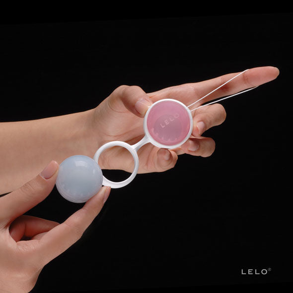 Main présentant les boules de geisha Luna Beads de Lelo sur fond noir, coque blanche avec anneau et sphères rose et bleue, accessoire d'entraînement périnéal.