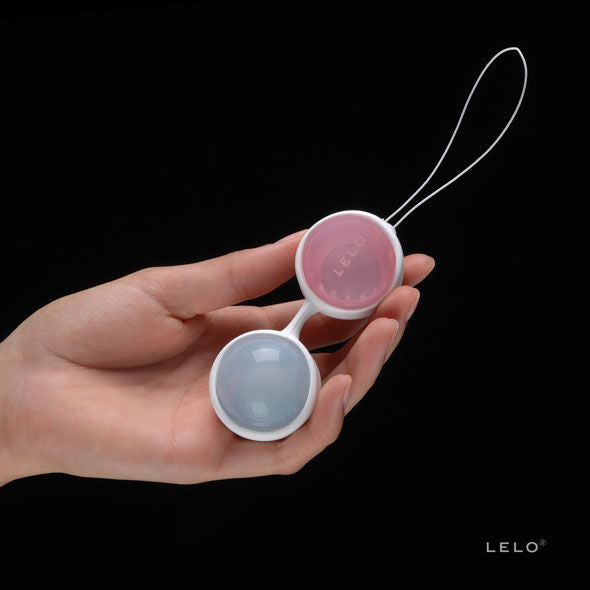 Main tenant les boules de geisha Luna Beads de Lelo assemblées sur fond noir, sphères rose et bleue avec cordon de retrait blanc, gros plan sur le set.